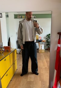 Lichtgrijze zip-up hoodie over een witte t-shirt met cropped hem, gecombineerd met losse zwarte broek; houten vloer en een potplant op de achtergrond.