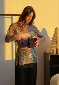 Femme portant un top à sequins scintillants et une jupe noire, tenant un sac à main noir, debout près d'un meuble en bois avec un arbre décoratif en papier sous une lumière chaude.