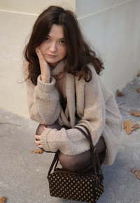 Beige knitted cardigan with ribbed texture, black handbag with gold eyelet accents, and dark tights. Model poses crouched against a wall.