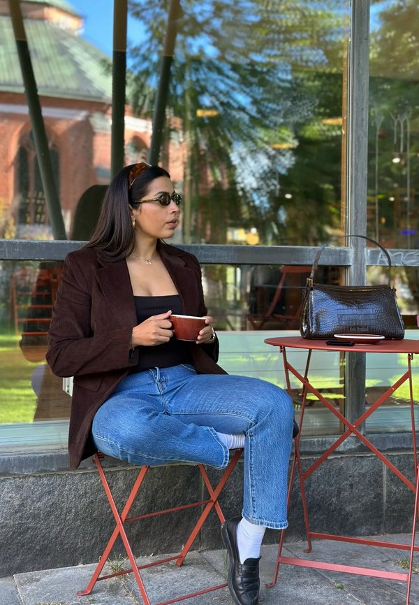 Mujer con gafas de sol y chaqueta marrón sentada con las piernas cruzadas en una silla naranja al aire libre, sosteniendo una taza cerca de una mesa a juego con un bolso.