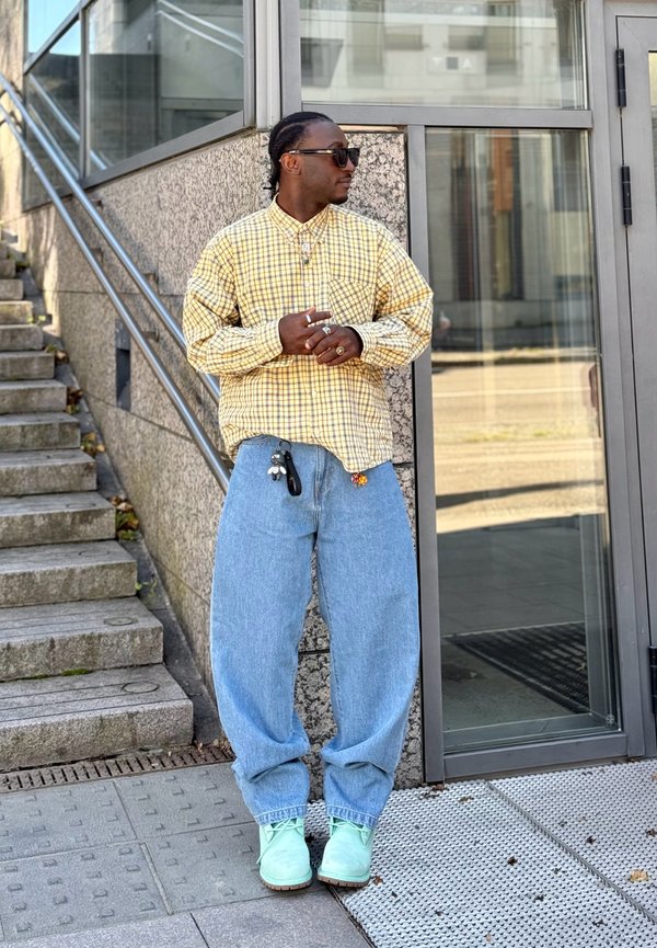 Yellow checkered long-sleeve shirt, baggy blue jeans, mint green shoes. Worn accessories include necklaces and multiple rings. Urban setting.