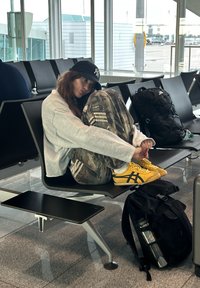 Hanorac gri confortabil, pantaloni camuflaj cu dungi, adidași galbeni Asics, relaxându-se pe un scaun de aeroport cu un rucsac negru aproape.