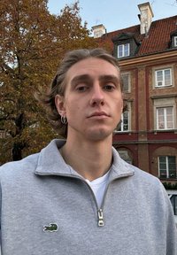 Grå fleecetop med lynlås og krave, med en lille grøn krokodille-logo. Baggrunden viser boligbygninger og efterårsblade.