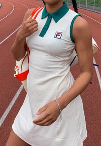 Robe blanche à rayures avec un col vert, ornée d'un petit logo rouge et bleu. Un sac blanc avec des accents rouges est porté en bandoulière.