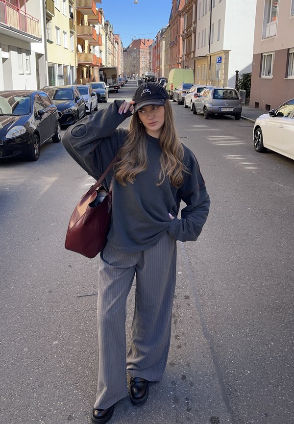 Suéter cinza oversized com detalhes em vermelho, calças largas cinzentas às riscas, boné preto e mala bordô. Sapatos de plataforma pretos. Cena de rua urbana.
