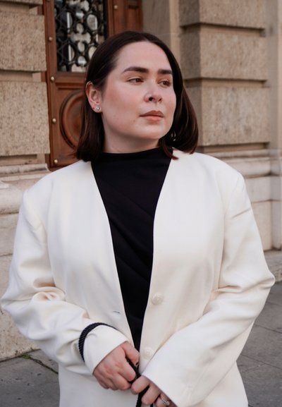 Mujer con cabello corto y oscuro, vestida con blazer blanco sobre top negro, de pie frente a un edificio de piedra con puerta de madera, mirando hacia la derecha.