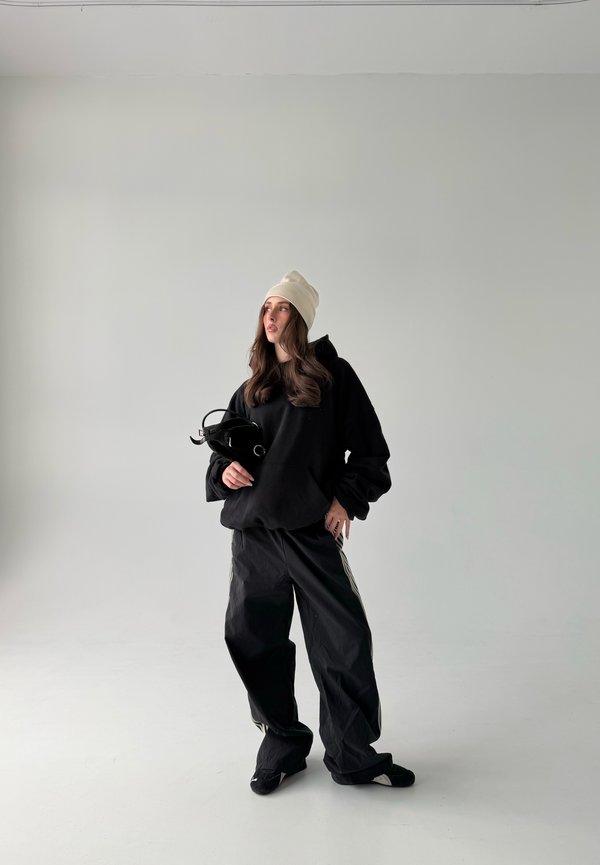 Kvinne iført svart oversized hettegenser, vide svarte bukser, beige lue, holder en liten svart veske, står foran ensfarget lysegrå bakgrunn.