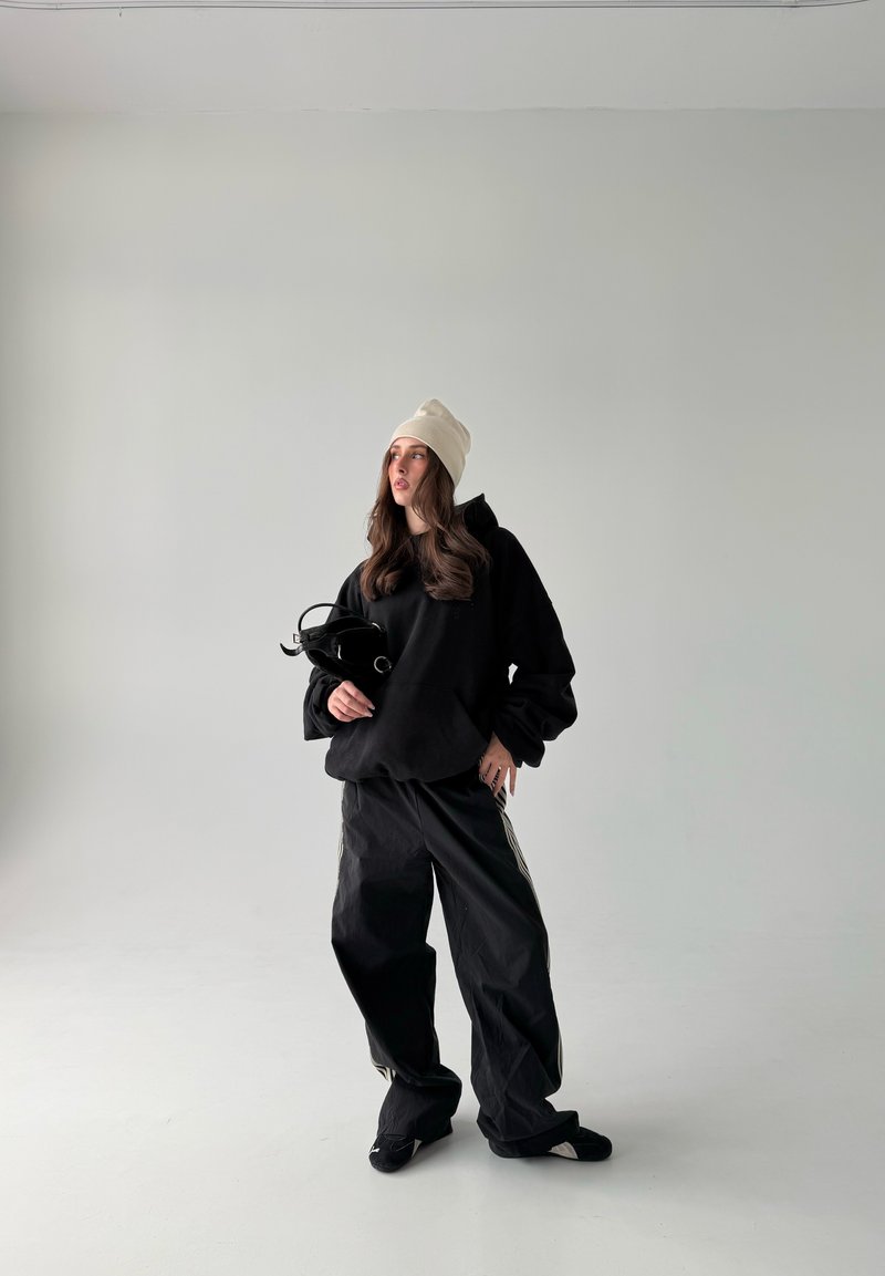 Nainen pukeutuneena mustaan ylisuurikokoiseen huppariin, leveisiin mustiin housuihin, beige pipo päässä, pitää kädessään pientä mustaa laukkua, seisoo yksivärisen vaaleanharmaan taustan edessä.