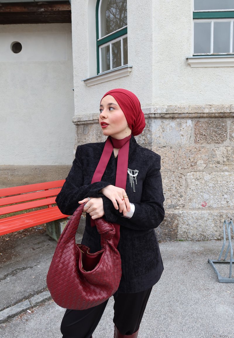 Femme portant un foulard rouge sur la tête, un blazer noir et une écharpe bordeaux, tenant un sac tissé bordeaux, debout à côté d'un banc rouge devant un bâtiment en pierre.