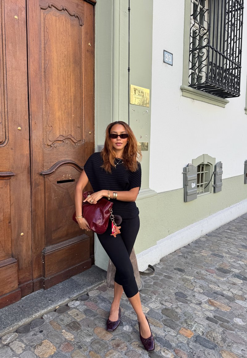 Una donna in un outfit nero e occhiali da sole si trova su una strada acciottolata, appoggiata a una grande porta di legno di un edificio bianco e verde.