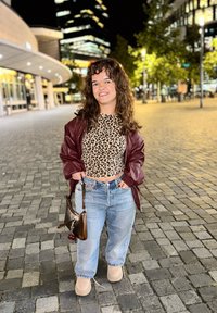 Person iført en leopardmønstret top, burgunder bomberjakke, lyseblå jeans med glitrende detaljer og beige sko. Holder en lille brun taske.