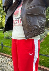 Brun jakke i kunstlæder med ribstrikkede manchetter, hvid t-shirt med grafisk print og røde joggingbukser med hvide sidelommer og Puma-logo.