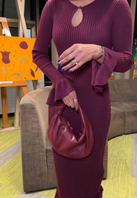 Άτομο που φοράει φόρεμα σε αποχρώσεις του μαρουν με ριγωτή υφή και φουσκωτά μανίκια, κρατώντας μια τσάντα σε ίδιο μαρουν χρώμα, με βαμμένα μπλε νύχια και πολλά δαχτυλίδια.