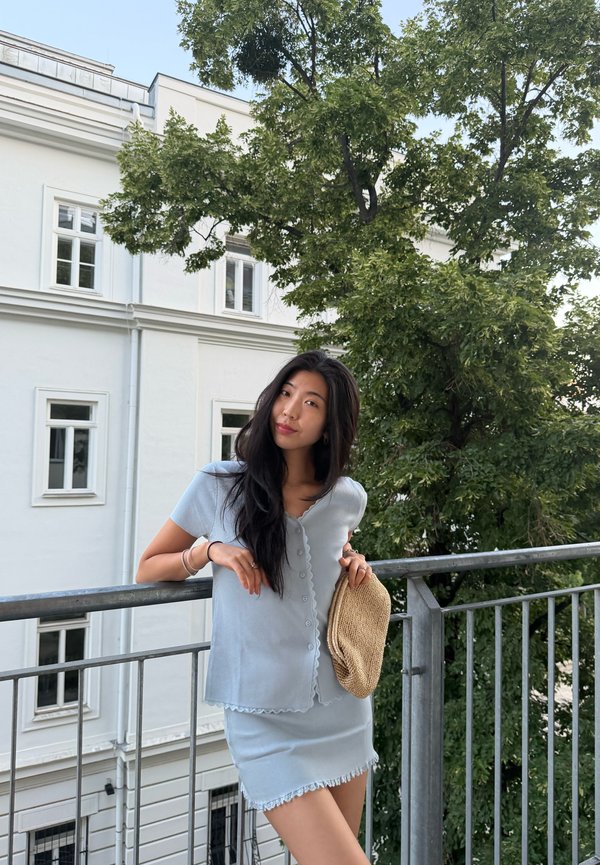 Giovane donna con lunghi capelli scuri si appoggia alla ringhiera del balcone tenendo un cappello intrecciato, indossa un top azzurro chiaro e una gonna, con un edificio e alberi sullo sfondo.