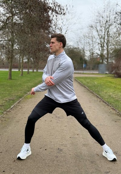 Atleta con una camiseta de manga larga gris claro, pantalones cortos negros y leggings de compresión negros, realizando una pose atlética en un sendero rodeado de árboles.