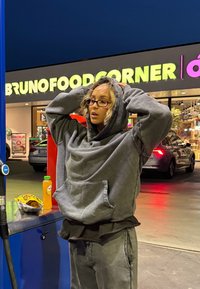 Felpa grigia oversize e pantaloni della tuta abbinati. L'individuo tiene il cappuccio e indossa occhiali. Vicino ci sono snack e una bevanda.