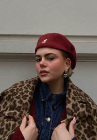 Jeune personne portant un béret rouge Kangol, un manteau en imprimé léopard, une veste en denim, des boucles d'oreilles en forme de cœur, et tenant le col du manteau contre un mur uni.
