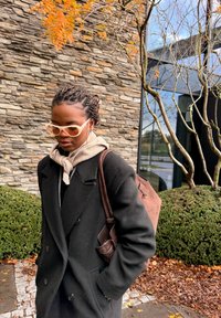 Cappotto nero doppiopetto, sciarpa beige annodata al collo, occhiali da sole oversize trasparenti, borsa a tracolla marrone, sfondo di muro di pietra testurizzato.