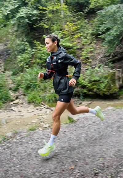 Chaqueta de running ligera negra con acentos naranjas, pantalones cortos negros y zapatos amarillos brillantes, ambientada en un entorno natural de senderos.