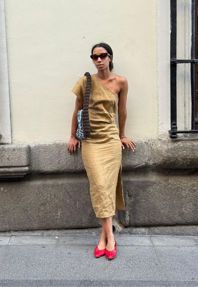 Vestido de lino marrón claro, con los hombros al descubierto, abertura lateral, combinado con un bolso de crochet marrón y zapatos planos rojos sobre una superficie de piedra texturizada.