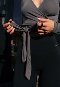 Femme nouant la ceinture d'un haut cache-cœur gris, portant des leggings Nike noirs, debout au soleil avec un fond sombre.