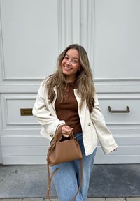 Creme dobbeltbrystet jakke over en brun ribbet top, holder en lille brun håndtaske. Blå jeans med løs pasform; glat finish.