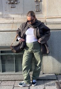 Kvinna som bär solglasögon, grå kappa och scarf, vit topp, gröna byxor och svarta sneakers, står mot en stenvägg och håller en brun handväska.