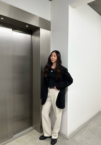 Mujer con cabello largo y ondulado, vistiendo un blazer y camisa negra, pantalones blancos, de pie junto a las puertas cerradas de un ascensor de acero inoxidable en un pasillo luminoso.