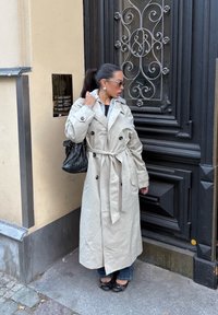 Femme avec une queue de cheval portant un long trench beige, des lunettes de soleil et un sac noir, se tenant près d'une porte noire ornée sur le trottoir de la ville.