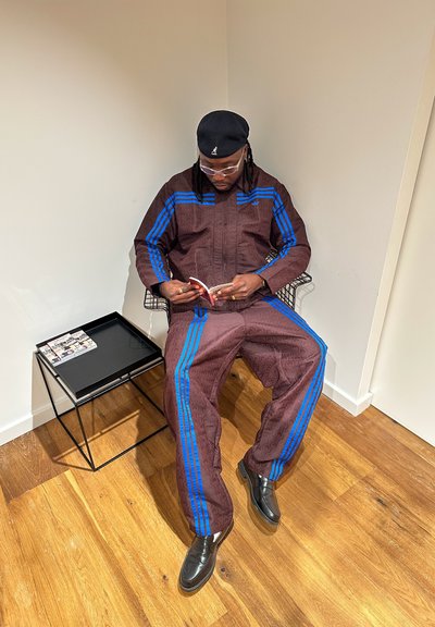 Chándal marrón con patrón y rayas azules, zapatos negros y gafas, sentado en una silla de alambre junto a una mesa auxiliar negra.