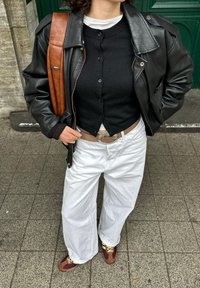 Persona vestida con una chaqueta de cuero negra, un cárdigan negro abotonado, pantalones blancos con un cinturón marrón, zapatillas de deporte burdeos y amarillas, y un bolso de hombro marrón.