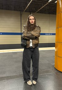 Smeđa kožna bomber jakna s rebrastim manšetama, crna torba, prevelike tamnosive traperice i svijetle cipele na stanici podzemne željeznice.