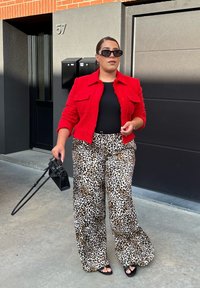 Rød tekstureret jakke over en sort top, parret med bredbenede bukser i leopardprint. Holder en lille sort håndtaske og bærer mørke solbriller.