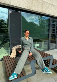Grønn ribbet zip-up genser med hvit krage, grå vidbukser og blå joggesko, sittende på en trebenk mot en glassbakgrunn.
