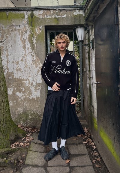 Persona joven con cabello rizado y rubio, vistiendo una blusa negra de manga larga y una falda larga negra, de pie en un pasaje estrecho al aire libre.