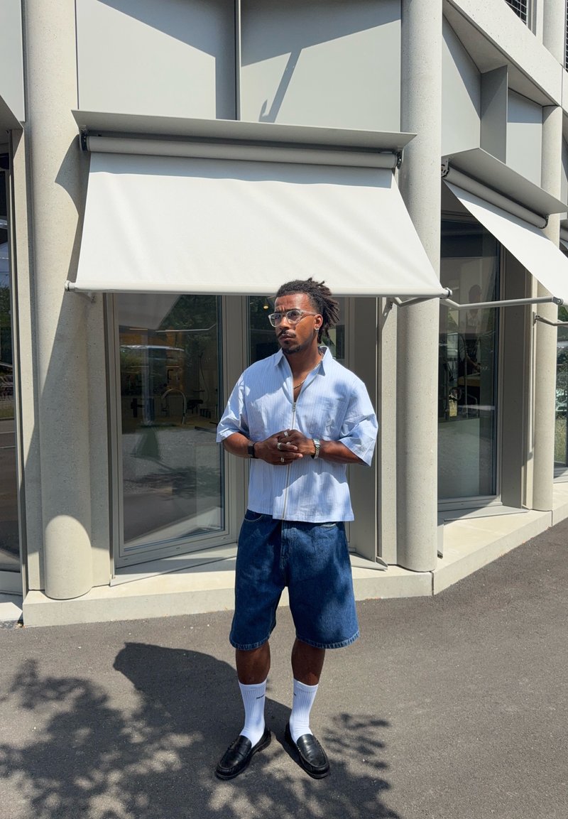 Uomo con occhiali che indossa una camicia azzurro chiaro, pantaloncini in denim, calzini bianchi e scarpe nere, in piedi all'aperto davanti a un edificio con tende bianche.