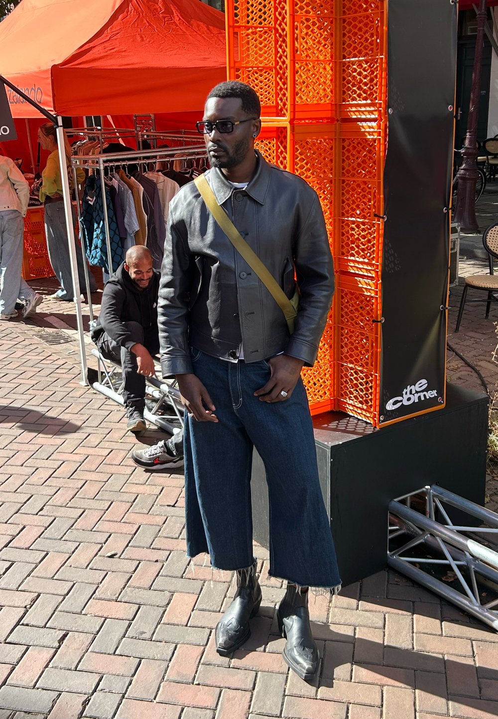 Sort læder cropped jakke med krave, denim culottes og sorte hælede støvler. Mand iført solbriller står mod en orange baggrund.