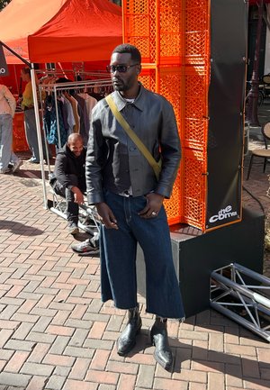 Juodas odinis trumpas švarkas su apykakle, džinsinės kelnės-culottes ir juodi kulniukai. Vyras, dėvintis akinius nuo saulės, stovi priešais oranžinį foną.