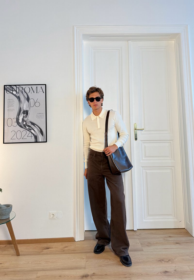 Pullover polo beige chiaro, pantaloni a gamba larga marrone scuro, scarpe nere e una borsa a tracolla nera, davanti a un muro bianco con un poster grafico.