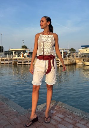 Top corto de encaje blanco, pantalones cortos blancos sueltos con ribete de encaje y un cinturón burdeos. Sandalias de cuña con acentos de madera marrón oscuro, en un entorno exterior junto al agua.