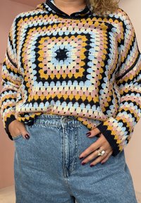 Sweatshirt de croché multicolor com um padrão quadrado em lã rosa, azul, amarelo e preto; combinado com calções de ganga de cintura alta.