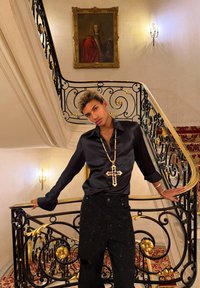 Jeune personne portant une chemise en satin noir et un collier croix perlée s'appuie sur la rampe d'escalier noire et dorée ornée, dans un intérieur élégant et bien éclairé.