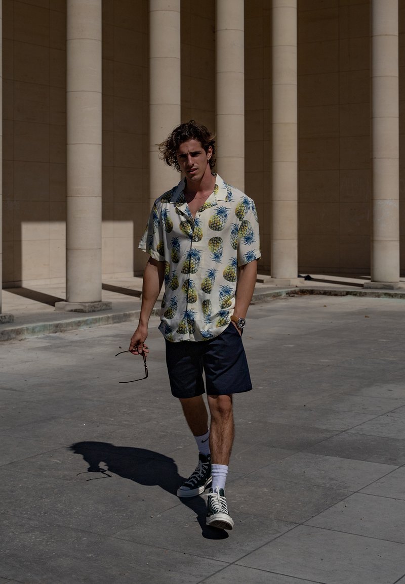 Trumpas rankovės marškinėliai su balta baze ir mėlyna ananasų printu, derinami su tamsiai mėlynomis šortais ir žaliais aukštais sportbačiais. Laiko saulės akinius.