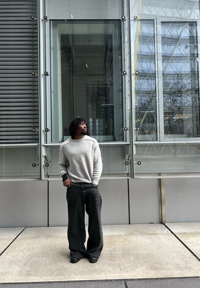 Joven con suéter gris y pantalones anchos negros, de pie con las manos en los bolsillos, mirando hacia arriba junto a la fachada moderna de un edificio de vidrio y metal.