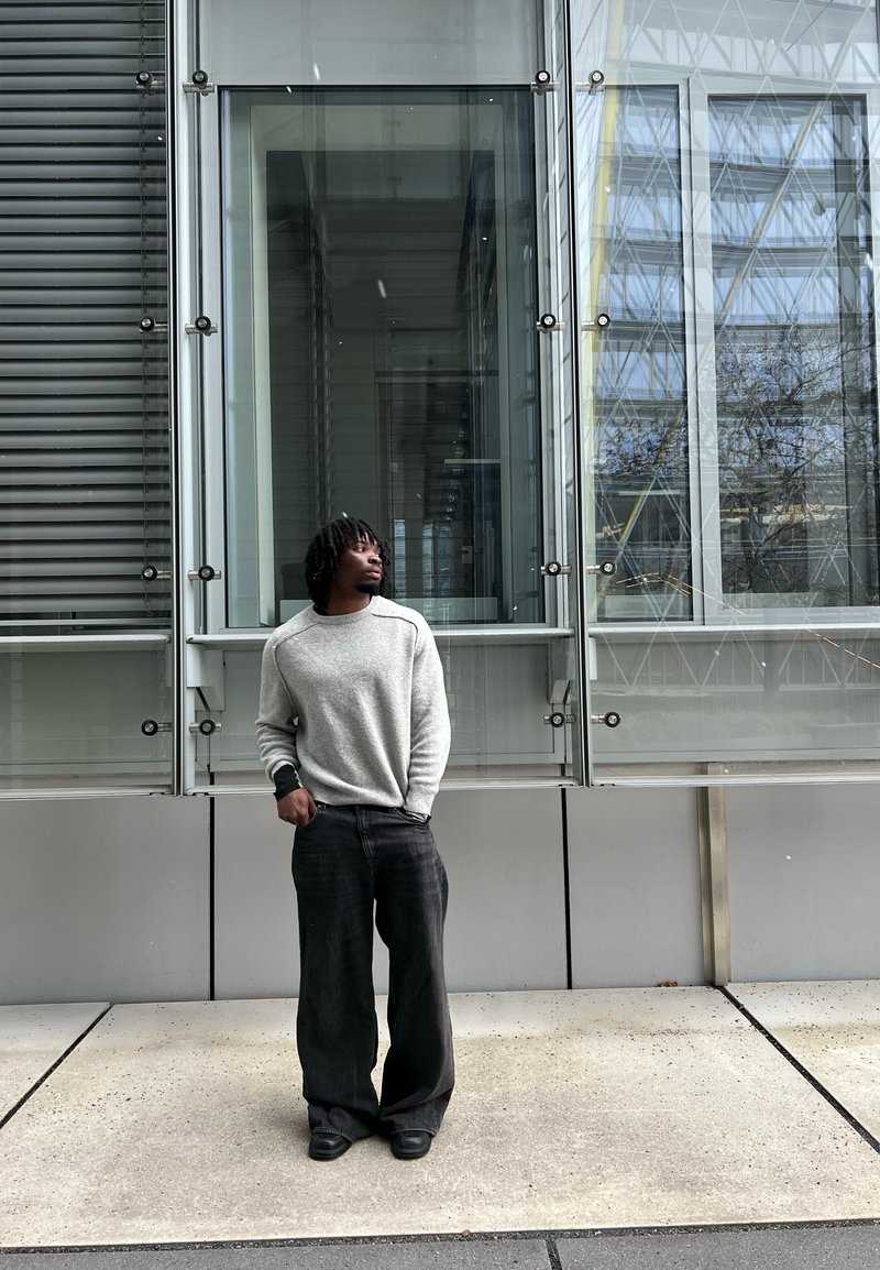 Jeune homme portant un pull gris et un pantalon large noir, debout les mains dans les poches, regardant vers le haut à côté d'une façade moderne en verre et métal.