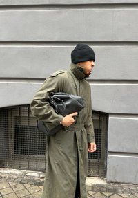 Ελιά πράσινο παλτό με ζωνάκι στη μέση, φορεμένο με ένα μαύρο σκουφί. Κρατώντας μια μαύρη δερμάτινη τσάντα messenger, περπατώντας σε έναν δρόμο.