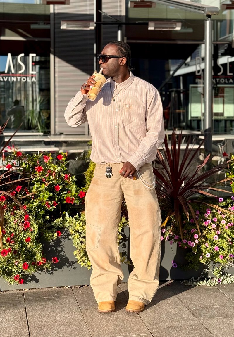 Pantalon en velours côtelé de couleur claire et ample, chemise à manches longues rayée, lunettes de soleil foncées, tenant un verre, sur un fond floral.