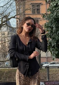 Femme portant des lunettes de soleil, blouse noire à volants, jupe imprimé léopard, debout à l'extérieur près d'arbres et d'un bâtiment en briques, ajustant ses cheveux.