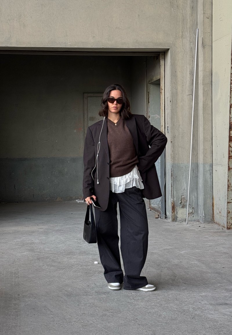 Kobieta w dużym ciemnym żakiecie, brązowym swetrze, białej bluzce z falbanami, szerokich czarnych spodniach, białych trampkach, w okularach przeciwsłonecznych, trzymająca czarną torebkę w przemysłowej przestrzeni.