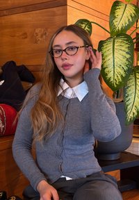 Jeune femme avec des lunettes et de longs cheveux, portant un cardigan gris et une chemise blanche, assise à l'intérieur à côté d'une grande plante en pot aux feuilles vertes.